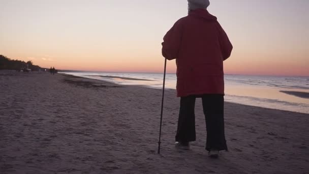 Літня жінка йде з палиці для нордичної ходьби вздовж берега моря. — стокове відео