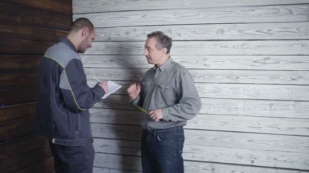 El cliente se fusiona con el constructor sobre la reparación de su habitación en el fondo de una pared de madera. El cliente quiere tener un armario — Vídeo de stock