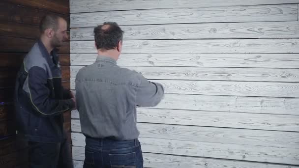 El cliente se fusiona con el constructor sobre la reparación de su habitación en el fondo de una pared de madera. El cliente quiere tener un armario — Vídeos de Stock