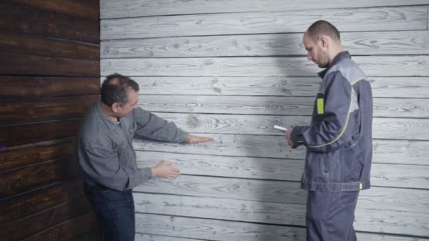 De klant wordt samengevoegd met de bouwer over de herstelling van zijn kamer op de achtergrond van een houten muur. De klant wil hebben een kledingkast — Stockvideo