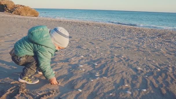 Estilo de vida do bebé. Um menino é jogado com areia e pedras em uma praia natural — Vídeo de Stock