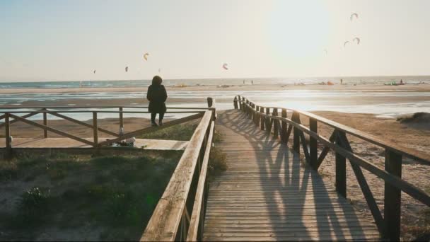 Un puente de madera, una mujer solitaria y muchos atletas practicando kitesurf en la playa del océano. Movimiento lento — Vídeos de Stock