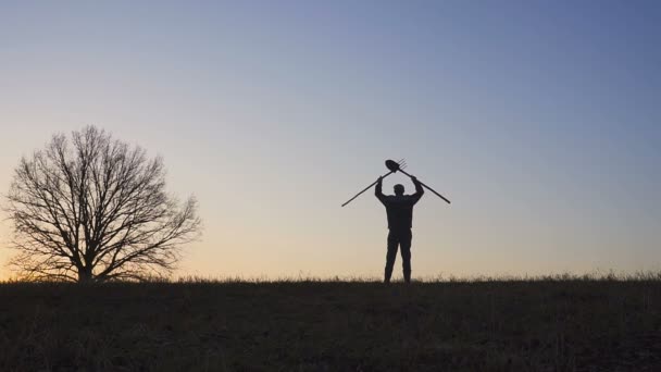 Un contadino soddisfatto che agita le mani con una pala e dei forconi. Silhouette di un tramonto o alba in campo . — Video Stock