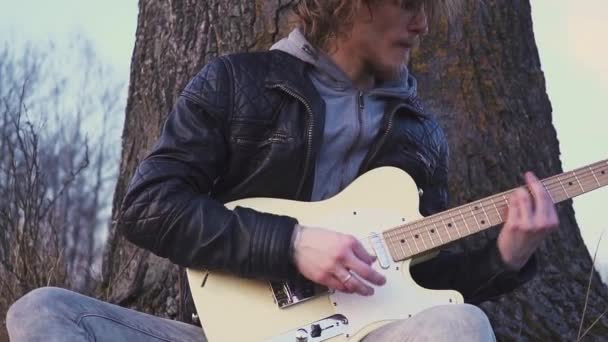 Hombre toca la guitarra eléctrica en un campo cerca del árbol al atardecer — Vídeo de stock