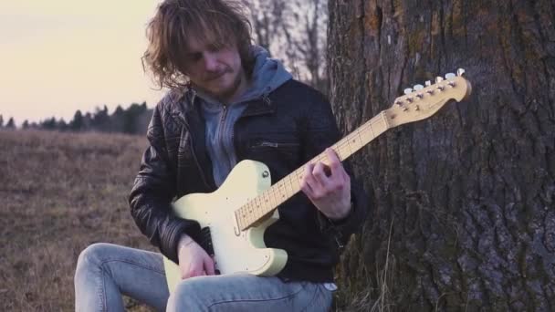 Hombre toca la guitarra eléctrica en un campo cerca del árbol al atardecer — Vídeos de Stock