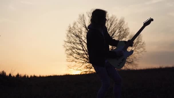 O homem toca guitarra elétrica e canta uma canção lírica em um campo perto da árvore ao pôr do sol. silhueta — Vídeo de Stock