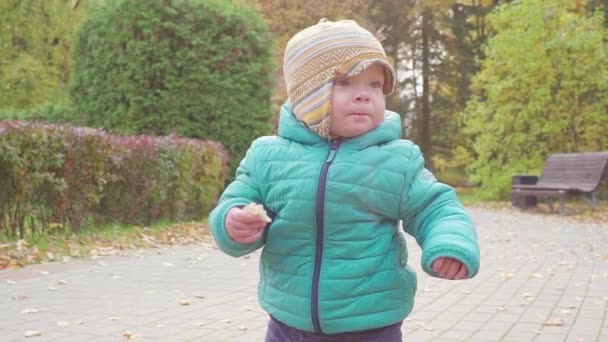 Lustig lächelnder 1-jähriger Junge, der im Herbstpark bei Sonnenuntergang mit Brot in der Hand spaziert. Glückliche Kindheit. Zeitlupe — Stockvideo