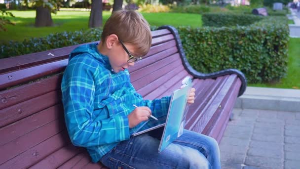 Portret van een schooljongen die huiswerk met een tekstboek en een tablet op het buitenleven doet — Stockvideo