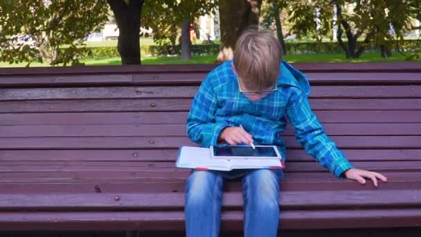 Retrato de un escolar que hace los deberes con un libro de texto y una tableta al aire libre — Vídeo de stock