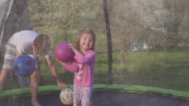 Frère et sœur jouant sur le terrain le soir. Ils sautent et jettent les balles dans la caméra, sautant sur le trampoline sur la pelouse. Concept de famille amicale. Mouvement lent — Video