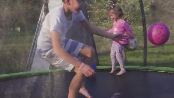 Brother and sister playing on the field at the evening time. People having fun outdoors. They jumping on trampoline on the lawn. Concept of friendly family. Spring. Slow motion — Stock Video