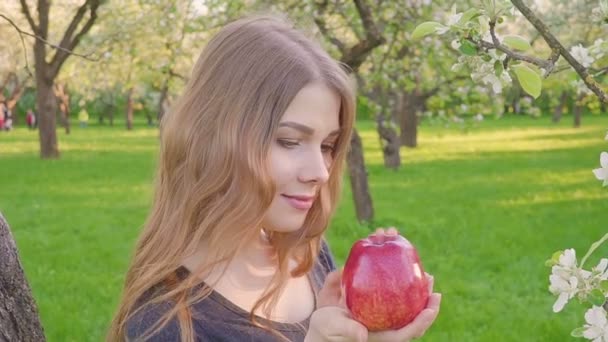 Schönes Mädchen mit einem Apfel in der Hand vor dem Hintergrund eines Apfelbaumgartens. eine Frau will einen Apfel essen. Garten mit blühendem Garten. Blumen von Obstbäumen. — Stockvideo