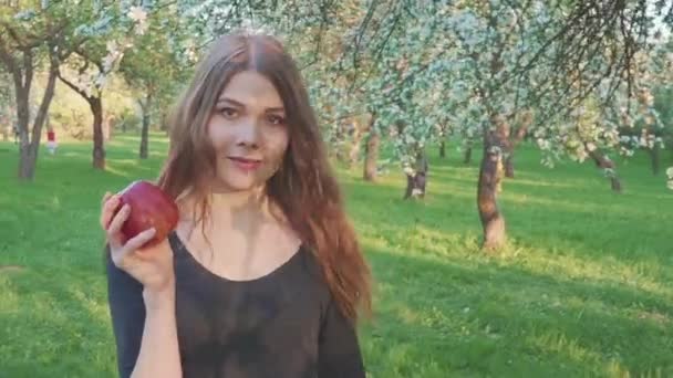 Schönes Mädchen mit einem Apfel in der Hand vor dem Hintergrund eines Apfelbaumgartens. eine Frau will einen Apfel essen. Garten mit blühendem Garten. Blumen von Obstbäumen. — Stockvideo