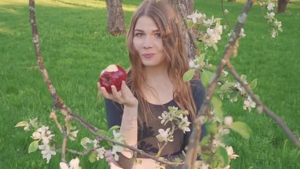 Mooie jonge vrouw met een appel in haar handen tegen de achtergrond van een appelboomgaard. Een vrouw eet een appel. Tuin met een bloeiende tuin. Bloemen van fruitbomen. — Stockvideo