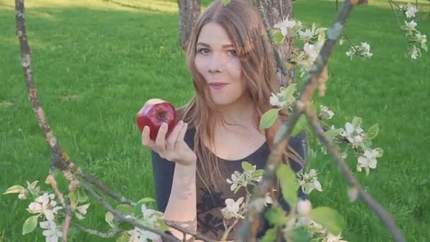 Krásná mladá žena s jablkem v ruce proti pozadí jablečného sadu. Žena jí jablko. Zahrada s kvetoucí zahradou. Květy ovocných stromů. — Stock video
