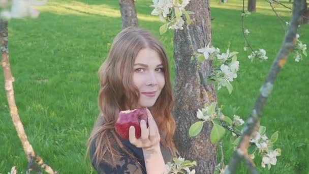 Frumoasă femeie tânără cu un măr în mâini pe fundalul unei livezi de mere. O femeie mănâncă un măr. Grădină cu o grădină înfloritoare. Flori de pomi fructiferi . — Videoclip de stoc
