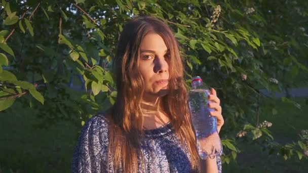 Hermosa excursionista bebiendo agua en el bosque al atardecer. Una chica en un vestido bebe agua fría de una botella de plástico . — Vídeos de Stock