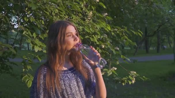 Beautiful female hiker drinking water in forest at sunset. A girl in a dress drinks cool water from a plastic bottle. — Stock Video