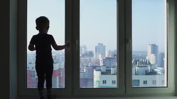 Силуэт маленького мальчика, стоящего у окна на фоне большого города с небоскребами — стоковое видео