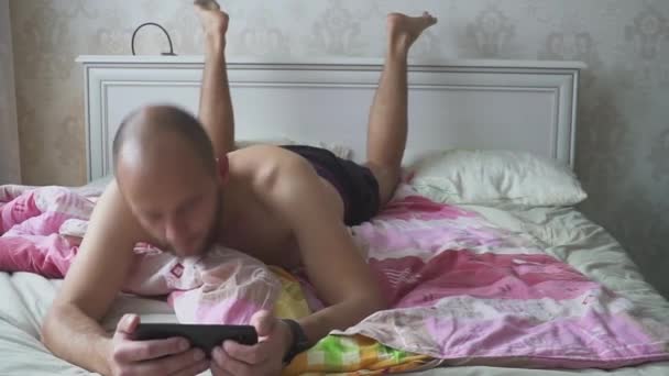 젊고 잘생긴 남자가 침대에서 스마트폰을 사용하는 모습. — 비디오