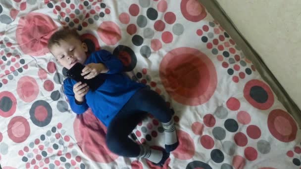 Niño acostado en la cama y mirando una pantalla de teléfono inteligente electrónico — Vídeos de Stock