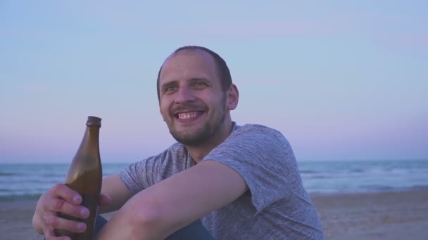 Пьяный мужчина пьет пиво на морском пляже вечером. Портрет человека, пьющего пиво из бутылки — стоковое видео
