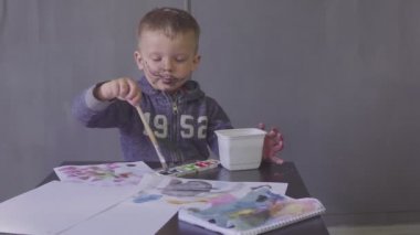 Kesik yüzlü komik bir çocuk renkli boyalarla resim çiziyor.