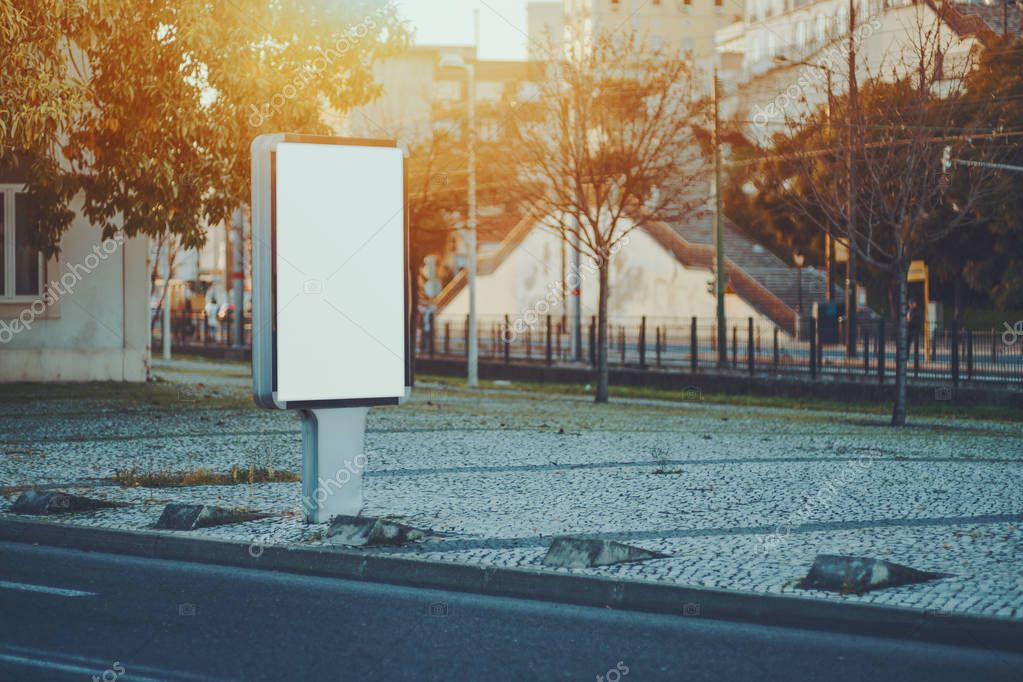 City blank information banner standing near road on paving stone, empty mock-up of urban poster next to highway, white clear billboard with copy space for logo, text or other advertising in evening