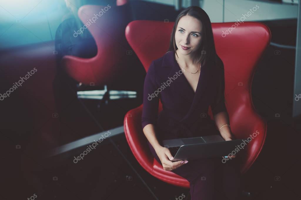 Portrait of adult pensive female entrepreneur wistfully looking aside while sitting in front of mirror on red bent armchair and holding laptop, with copy space zone for text, your logo or advertising