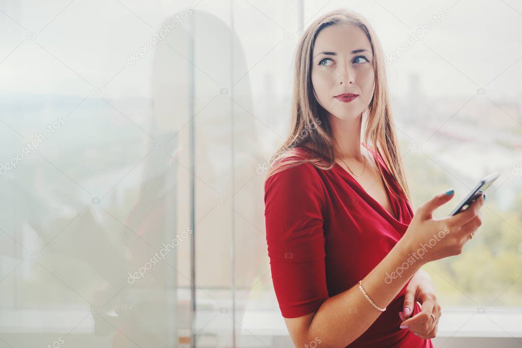 Businesswoman in red dress with smartphone near office window