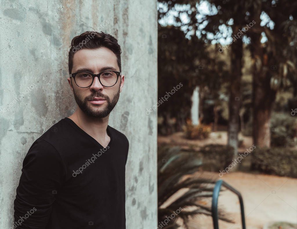 Portrait of the hipster bearded guy in glasses outdoors leaning against a concrete wall; the handsome young bearded man next to a cement wall with copy space place for your advertising, logo or text