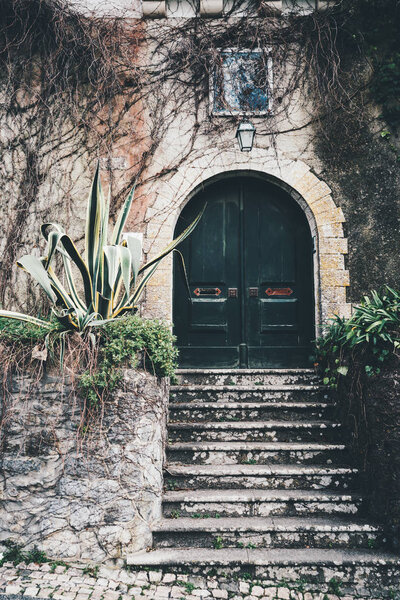 An antique wooden gate in the recess of a stone arch in the wall with a flaked plaster, stony stairs in front, a small lantern on the top surrounded with dry branches, Sintra, Portugal