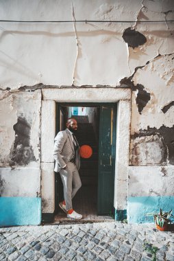 Süslü, süslü, sakallı bir Afrikalı adam elinde bir basketbol topuyla kapıda duruyor; siyah zarif adam kapının önünde bir ekoseli kostüm kazanıyor ve dışarıda sarkık bir duvar ve koyu bir koridor var.