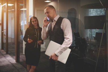 Two business partners are waiting for the beginning of a meeting in an office corridor near the entrance to a board room; selective focus on the woman entrepreneur, her colleague is phoning near her