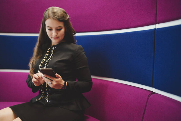 A charming mature hispanic woman in a formal suit is reading an e-mail on the screen of her cellphone while sitting on a bent lilac and classic blue sofa with a copy space place on the right