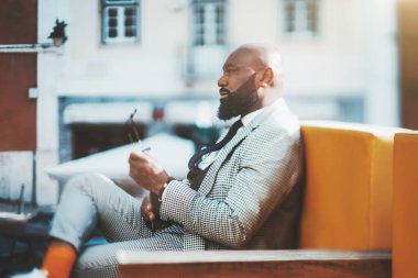Düzgün sakallı, bakımlı bir kostüm ve turuncu çoraplı, güneşli bir günde, açık havada koltukta oturan, yüzü seçici bir şekilde seçilmiş kel bir Afrikalı 'nın gerçek portresi.