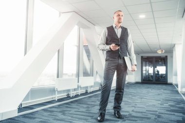 Uzun boylu, yakışıklı, zarif, beyaz bir girişimci elinde akıllı bir telefon ve dizüstü bilgisayarla parlak bir koridorda duruyor, modern bir ofis geçidinde görkemli bir İspanyol işadamı.