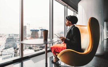 A ravishing African-American businesswoman on a yellow armchair is using her tablet PC; biracial woman entrepreneur using a digital tablet being on the top floor of a luxury business office skyscraper clipart