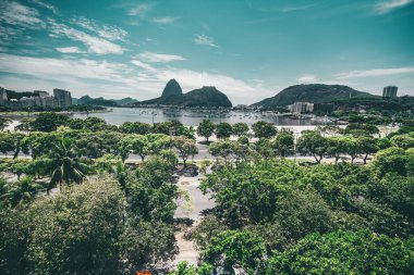 Rio de Janeiro, Brezilya 'nın Botafogo bölgesinin geniş açılı görüntüsü. Önünde birden fazla palmiye ve diğer tropikal ağaçlar var. Uzakta yelkenli tekneleri ve Sugar Loaf dağı olan bir körfez.
