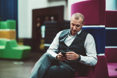 Yakışıklı, olgun, beyaz bir işadamı, modern ofis dinlenme alanının içindeki leylak kıvrımlı bir kanepede otururken akıllı telefonunda gelen bir mesajla şaşırtır.