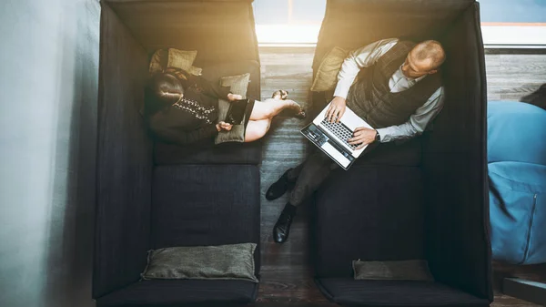 A businesswoman with a tablet pc and a man entrepreneur with a laptop in a coworking area indoors of a modern office are sitting on a soundproof sofa and having a leisurely business conversation