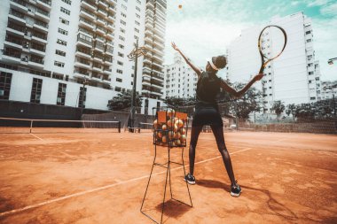 Spor antrenmanı sırasında tenis topuna ya da tenis kortunda ısınma antrenmanına vurmadan önce vurmayı hedefleyen zayıf, çift ırklı genç bir kadının geniş açılı görüntüsü, yanında topları olan metal bir sepet.