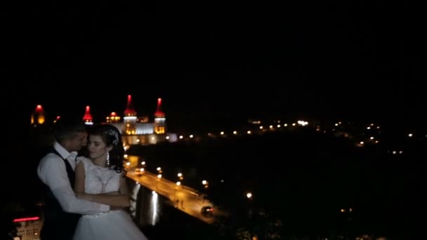 Piękny zamek na wzgórzu. Ufortyfikowany Pałac na skałach w nocy. Wieże z czerwono niebieskie podświetlenie. Para całuje się na tle zamku. — Wideo stockowe
