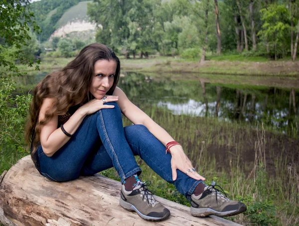 Giovane donna con i capelli lunghi in jeans seduta su un tronco pensieroso sulla riva di un piccolo lago forestale su uno sfondo di montagne e alberi in lontananza Foto Stock
