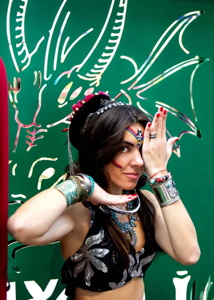 Young woman in ethnic jewelry and oriental clothes performing a dance gesture covering one eye by hand on the background colorful carved scenery