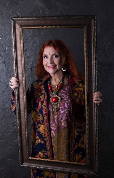 Redhead woman in ethnic jewelry and embroidered oriental coat fun laughs holding a picture frame on background the dark walls