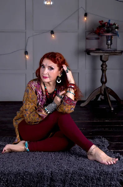 Redhaired woman in ethnic jewelry and oriental clothes sitting cross-legged propped on hand to head on the background wall with bulbs and a table with cells
