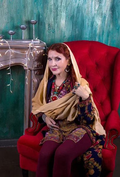 Vörös hajú nő indiai öltözékben és etnikai ékszer ül egy piros egy karosszék, a háttérben a zöld falakat és kandalló él — Stock Fotó