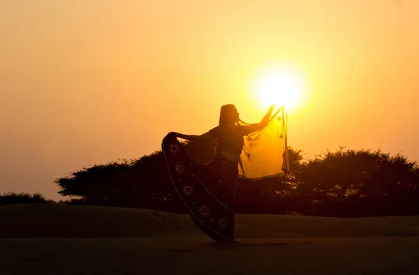 Una chica vestida de india baila al atardecer en el desierto en las dunas levantando la mano al sol contra los arbustos bajos y el cielo. Rajastán, India . — Foto de Stock