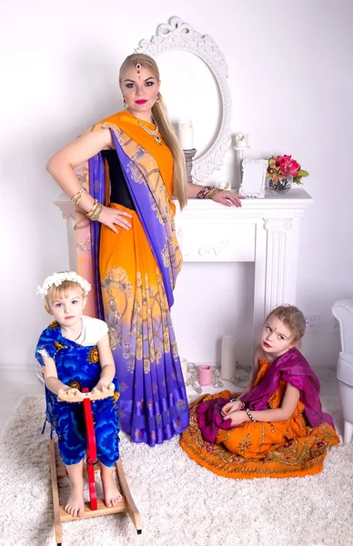 Joven mujer rubia en un brillante saris indio y joyas se encuentra junto a una chimenea blanca, y su hija pequeña sentada juntos en el suelo y caballo de madera —  Fotos de Stock